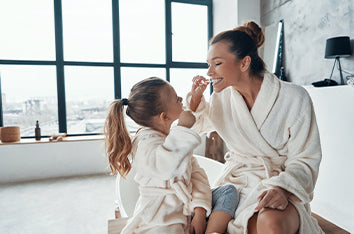 A mom and daughter in white bathrobes sit in the bathroom