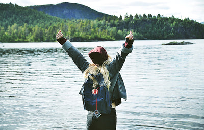A woman with her back to the camera raises her arms while facing a lake