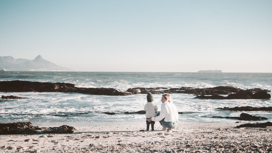 A woman and toddler boy stand on the beach by the ocean